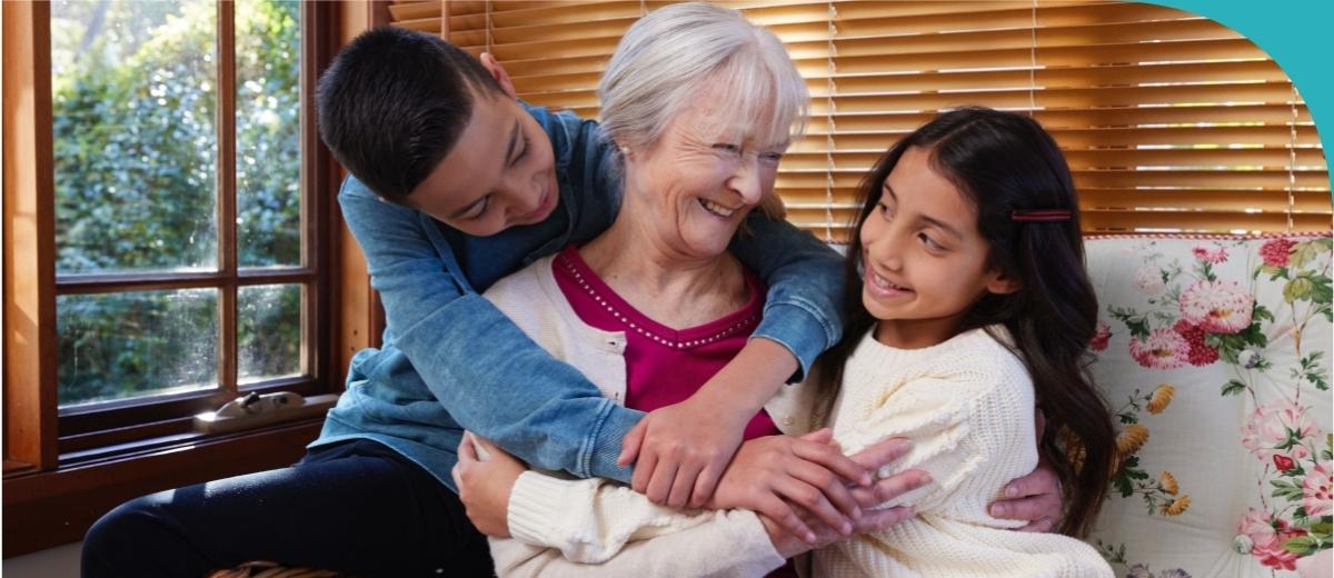 An older woman sits on a couch with a child on either side, hugging her. They smile at each other.