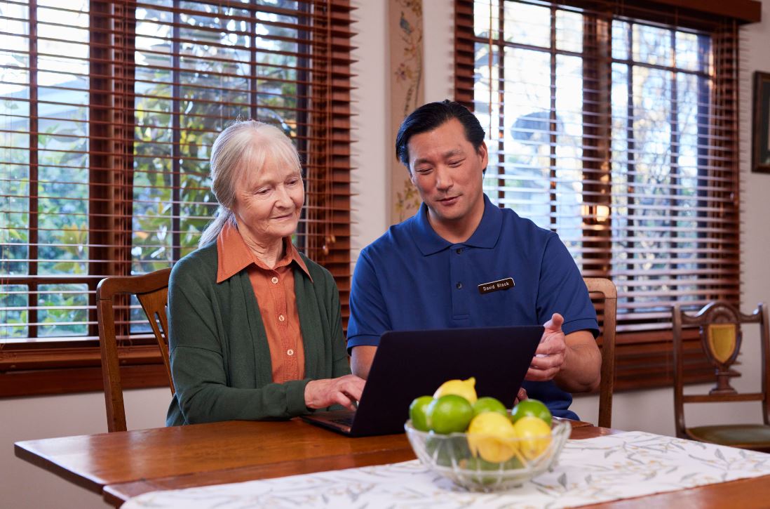 Older lady typing of a laptop with a support worker helping. Bowl of fruit in the foreground