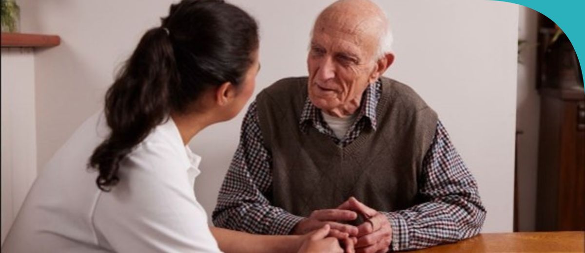 A healthcare professional, in a white uniform, sits facing an older man. They are in a homely setting, with the man dressed in a sweater and plaid shirt. They are holding hands across a table, suggesting a moment of care or comfort.