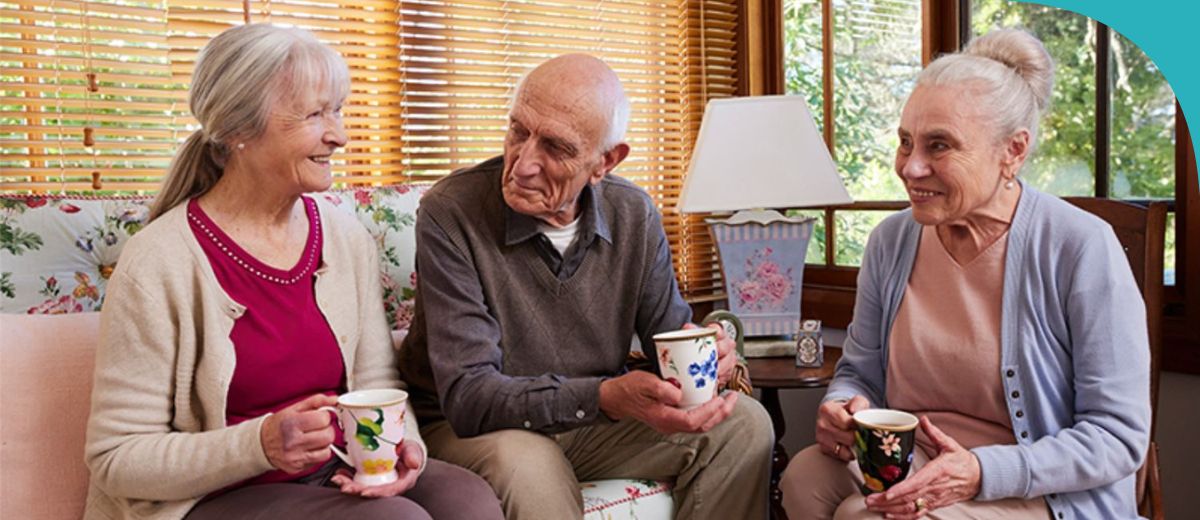 Three older people are seated in a warmly lit living room with a traditional decor. On the left, a woman smiles while holding a floral mug, in the center, an older man holds a similar mug with a somewhat somber expression, and on the right, another woman, also with a mug, is smiling gently. They appear to be enjoying a friendly conversation over tea or coffee.