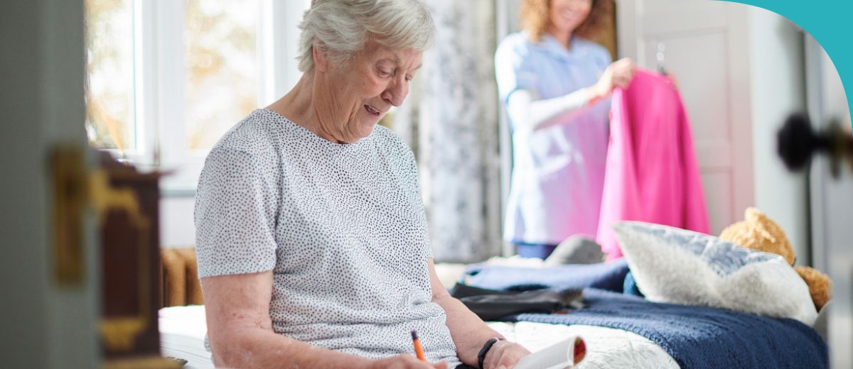 An older woman with short grey hair, sitting on a bed and smiling as she writes in a notebook. She is wearing a white and black polka dot top. In the background, a caregiver in a blue outfit is holding up a bright pink garment, possibly suggesting a choice of clothing.