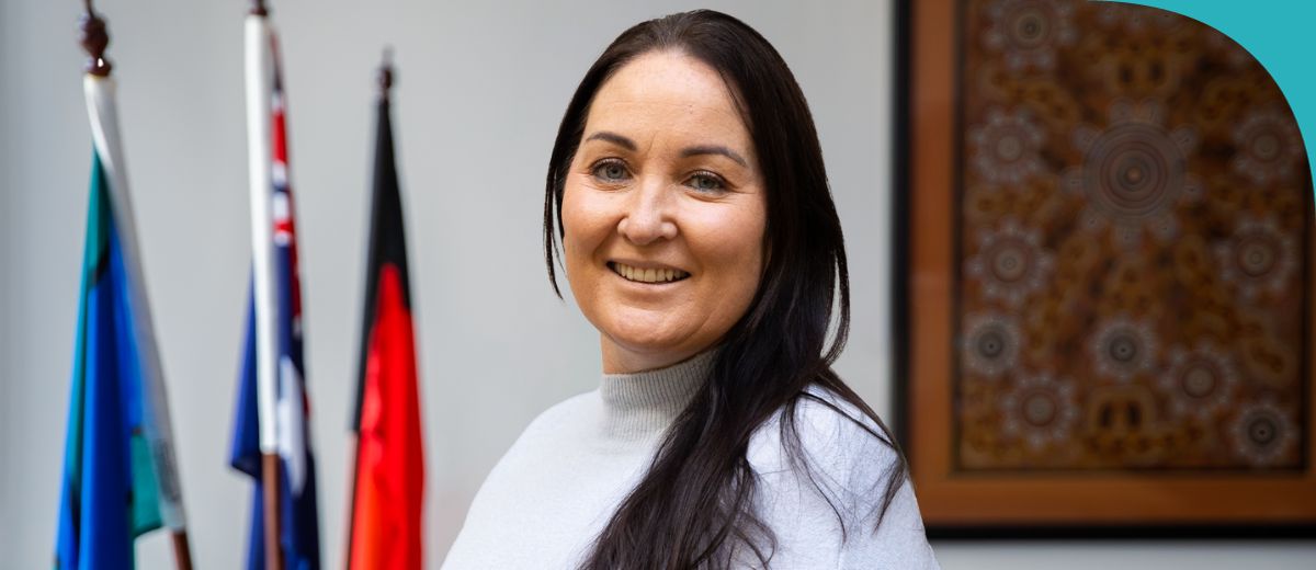 A woman in the foreground, smiling at the camera. She has long dark hair and is wearing a light-coloured turtleneck. In the background, there are several flags displayed on flagpoles. There is also a framed piece of artwork that seems to be of Aboriginal design, with circular patterns and a warm, earthy colour palette.