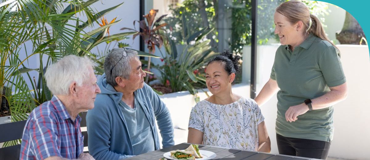 A group of three people are engaging in conversation at an outdoor table setting, with a waitress standing beside the table, smiling as she interacts with them. The setting appears relaxed and is likely at a café or a casual dining area, surrounded by green plants.