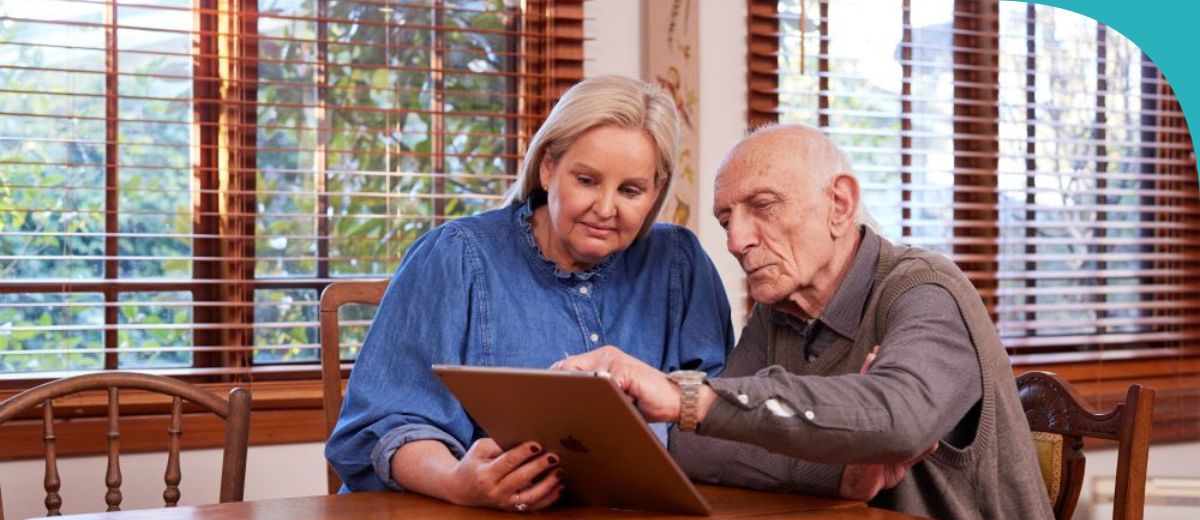 An older gentleman and a middle-aged woman are sitting at a wooden dining table indoors, with the woman showing the man something on a tablet. The room is bright with natural light coming through Venetian blinds. The scene suggests a home environment and could imply a family member assisting with technology.