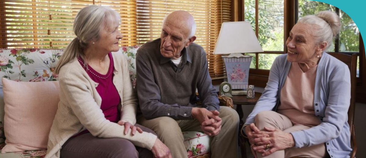 Three older individuals are engaged in a conversation while sitting in a living room with a warm atmosphere. Blinds cover the windows, and the room is decorated with traditional furniture and a floral patterned sofa.