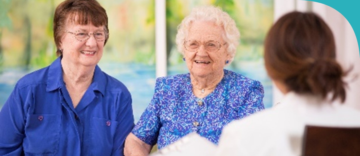 Two older women are smiling and conversing with an out-of-focus person across from them, likely in a healthcare or social setting.