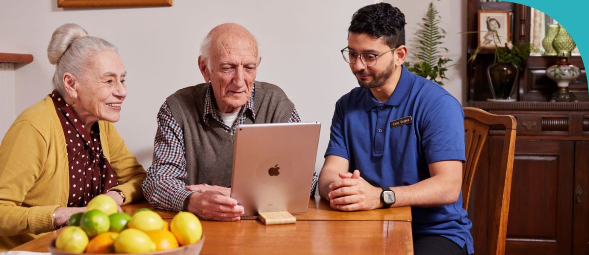 The image shows three individuals around a table with a bowl of citrus fruits in the foreground. An older man and woman are looking at a tablet screen, showing interest and engagement, while a younger man in a blue polo shirt, presumably a tech support worker given his embroidered badge, is explaining or assisting them.