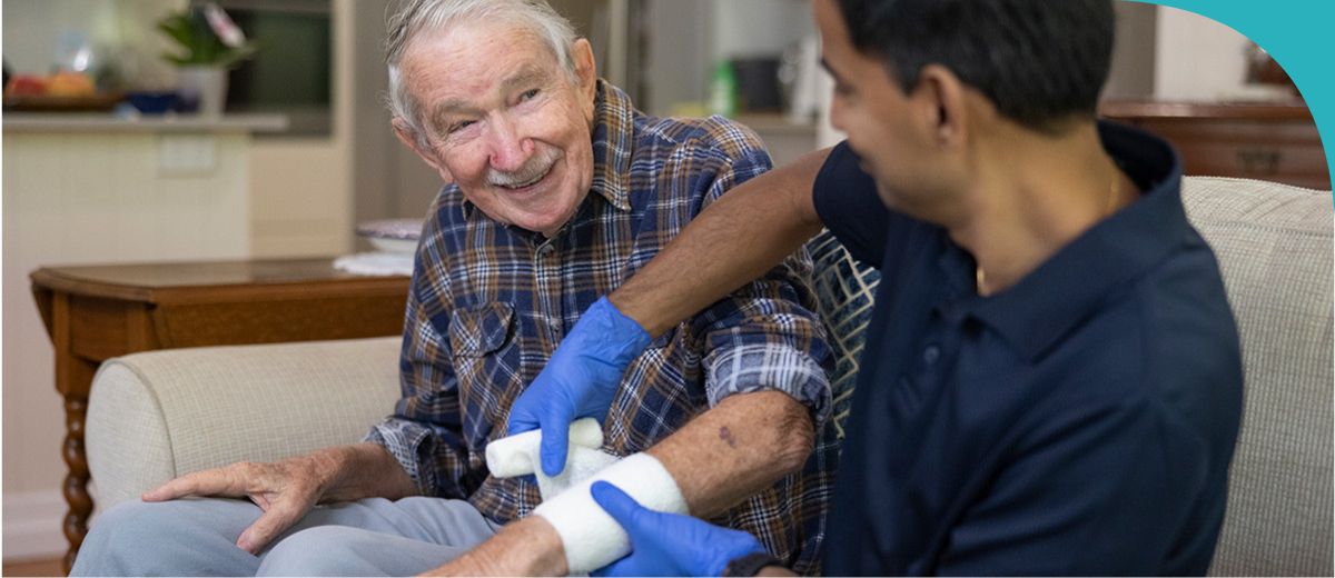 An older man and younger man sit next to each other on a couch. The younger man wears latex gloves and is wrapping a bandage around the older man’s forearm. The older man smiles at the younger man.