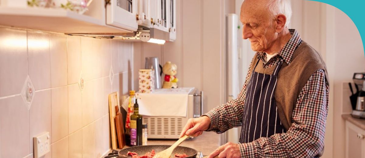 An elderly man is cooking in a kitchen, stirring a pan with his right hand. He is wearing a checkered shirt, a dark vest, and a striped apron. The kitchen is well-lit and appears homey, with white tiles and some kitchen items visible in the background.