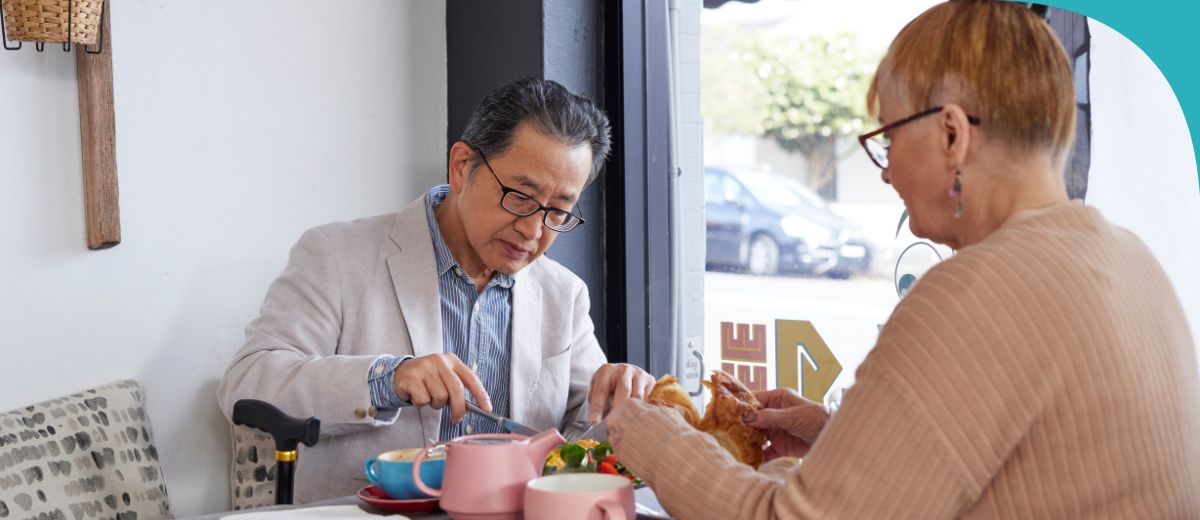 A man and woman, both seniors, sit at a table in a café eating food.