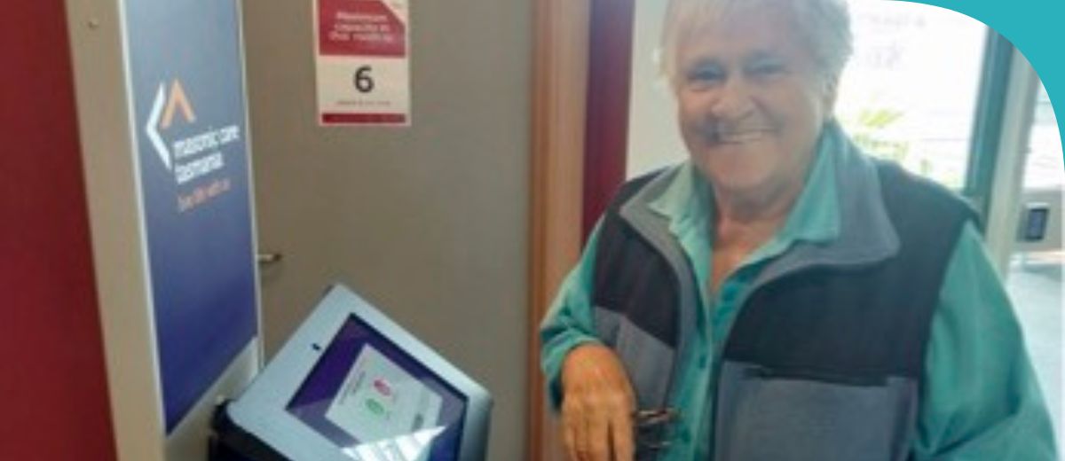 An older woman stands near a doorway in from of a touch screen station, smiling at the camera.