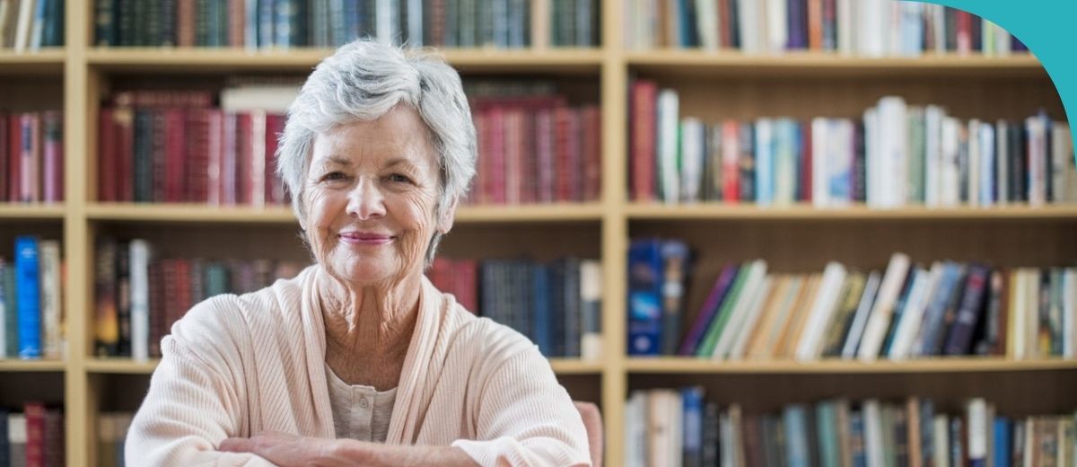 A smiling older lady with short white hair stands in front of wooden bookshelves filled with various books. She is wearing a soft pink cardigan and appears contented and relaxed.