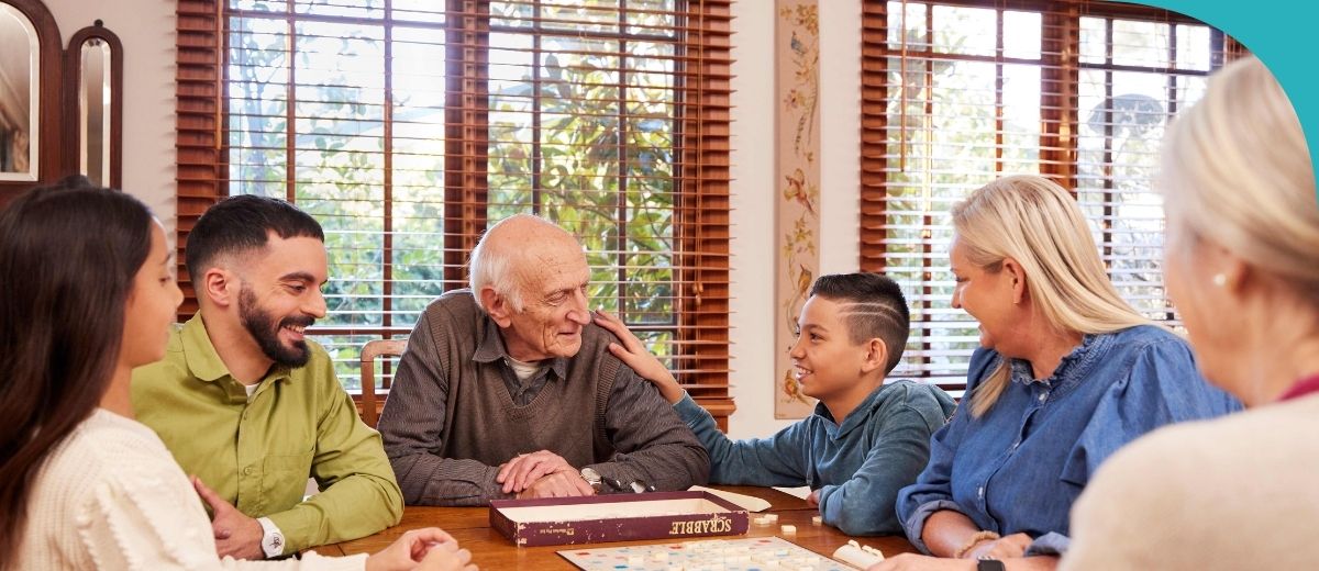 A family gathers around a dining table, engrossed in conversation and a board game. An older man shares a warm moment with a young boy, while others smile and engage with each other.