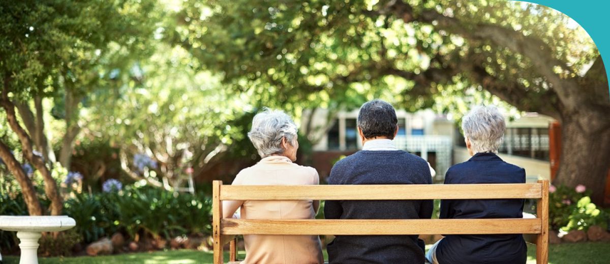 Three senior individuals are seated side by side on a wooden bench, facing a lush garden with dense foliage in a tranquil outdoor setting. They appear to be enjoying a quiet moment together in the serene surroundings, indicative of companionship and reflection.