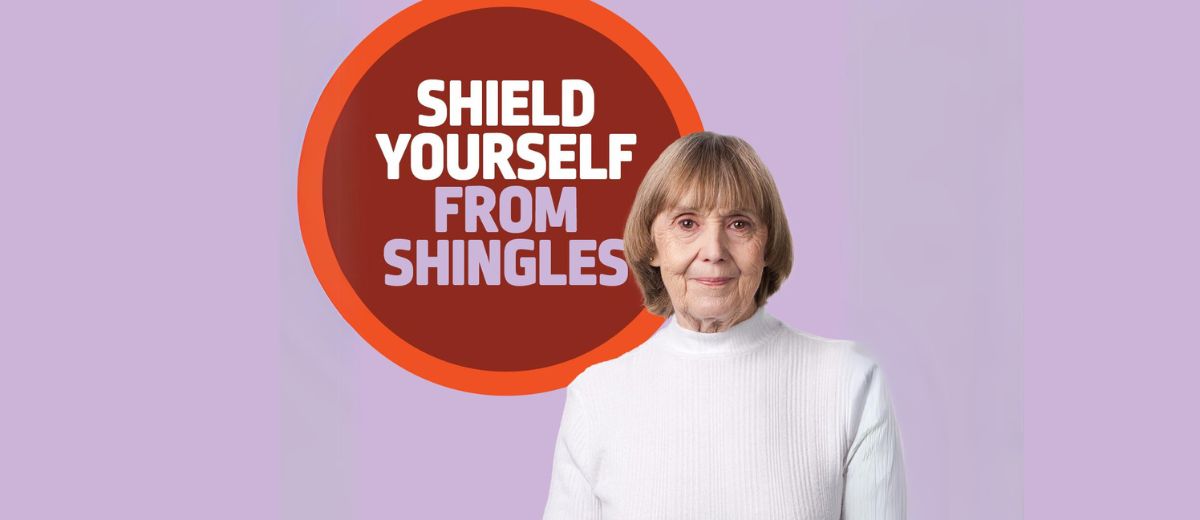 An older woman with short hair and a white turtleneck looks directly at the camera against a gradient purple background with a prominent orange circle that reads SHIELD YOURSELF FROM SHINGLES.
