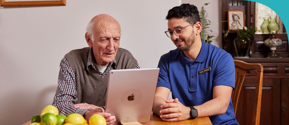 An older man and a younger man are sitting at a wooden table, looking at a tablet screen together. The table has a bowl of fresh citrus fruits, and the setting appears to be a cozy home interior